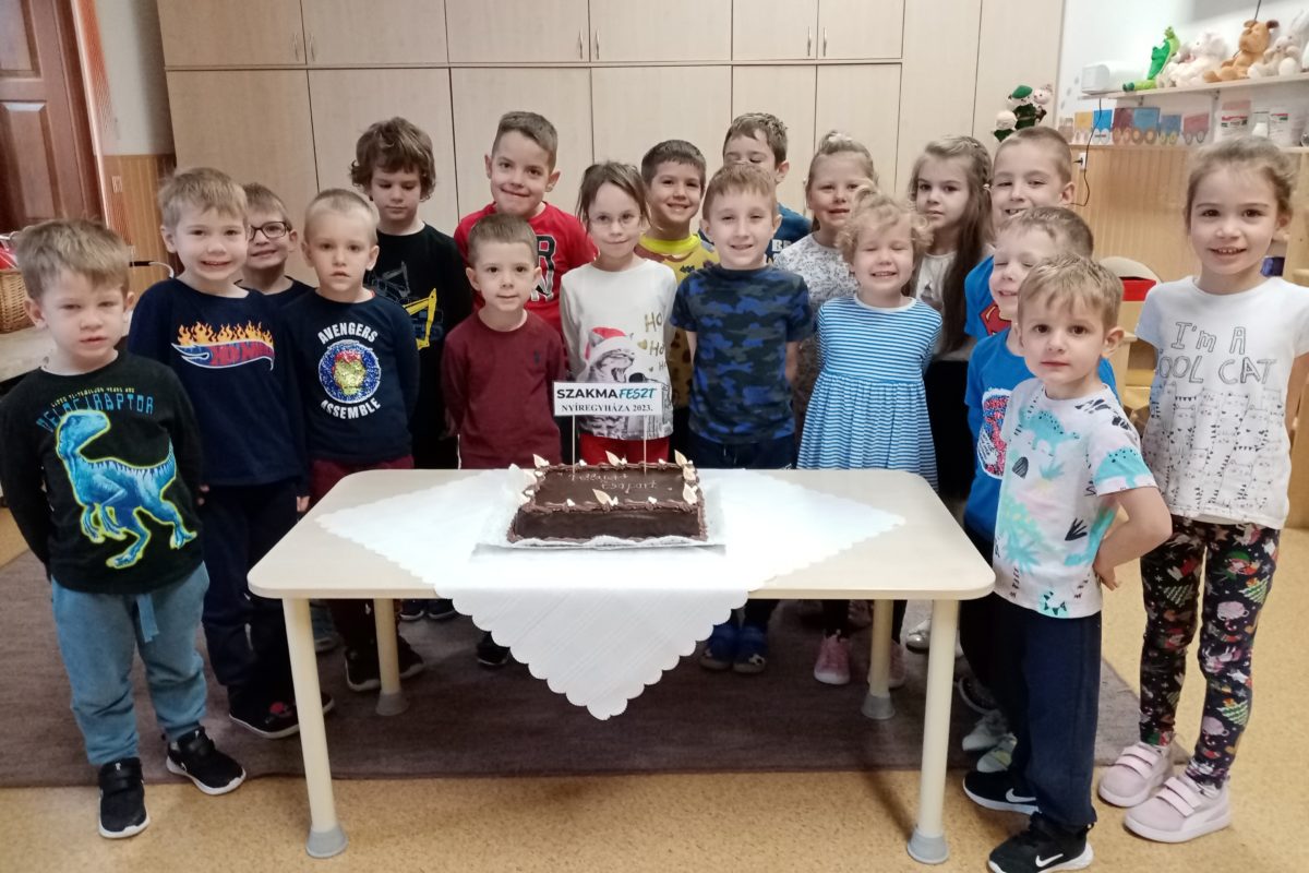 Egy csoport gyerek áll egy asztal körül, rajta egy tortával. A torta téglalap alakú, csokoládémázzal és gyertyákkal. A gyerekek mosolyogva nézik a kamerát. A szobában fából készült szekrények és polcok vannak játékokkal a háttérben.