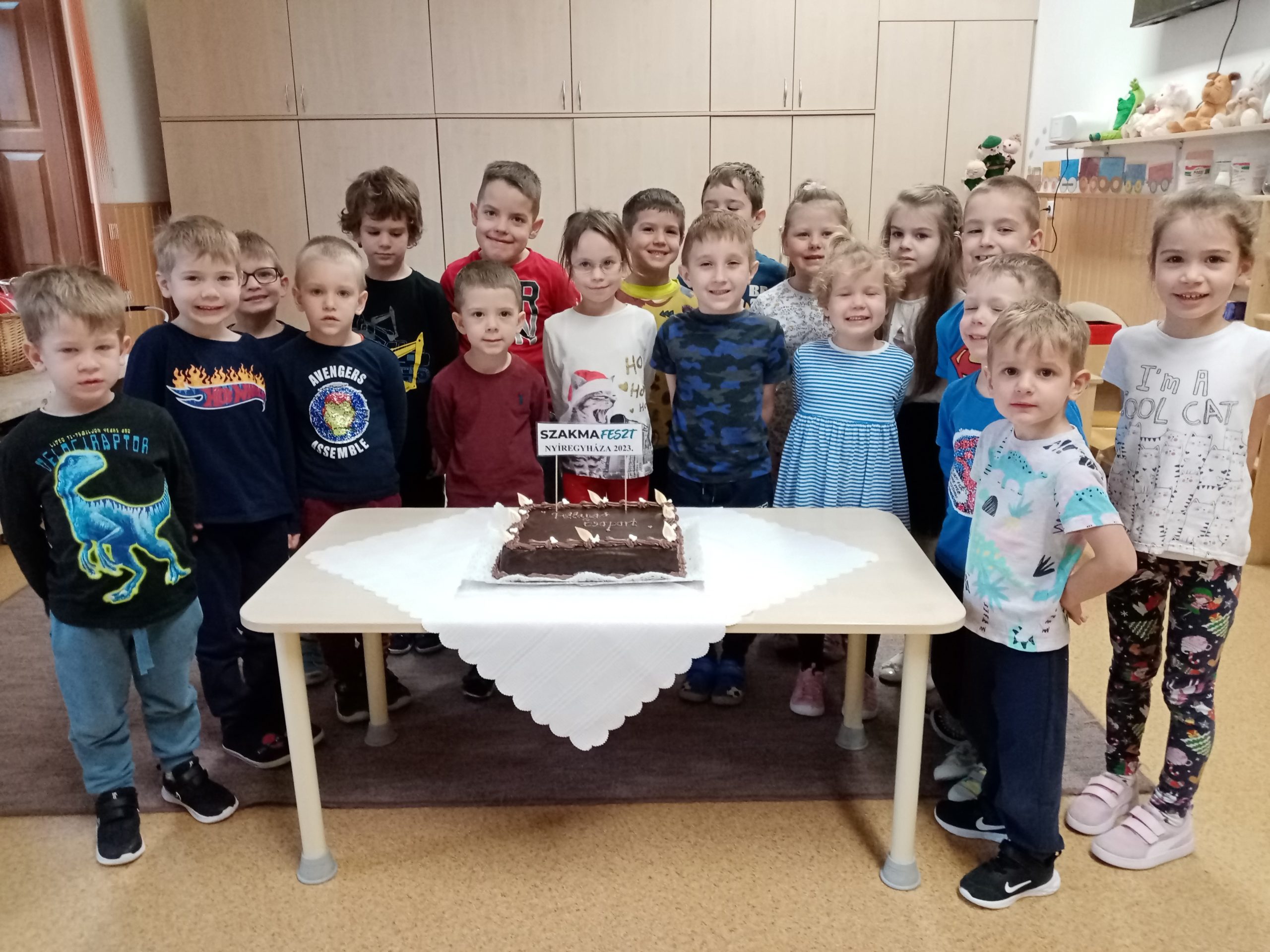 Egy csoport gyerek áll egy asztal körül, rajta egy tortával. A torta téglalap alakú, csokoládémázzal és gyertyákkal. A gyerekek mosolyogva nézik a kamerát. A szobában fából készült szekrények és polcok vannak játékokkal a háttérben.