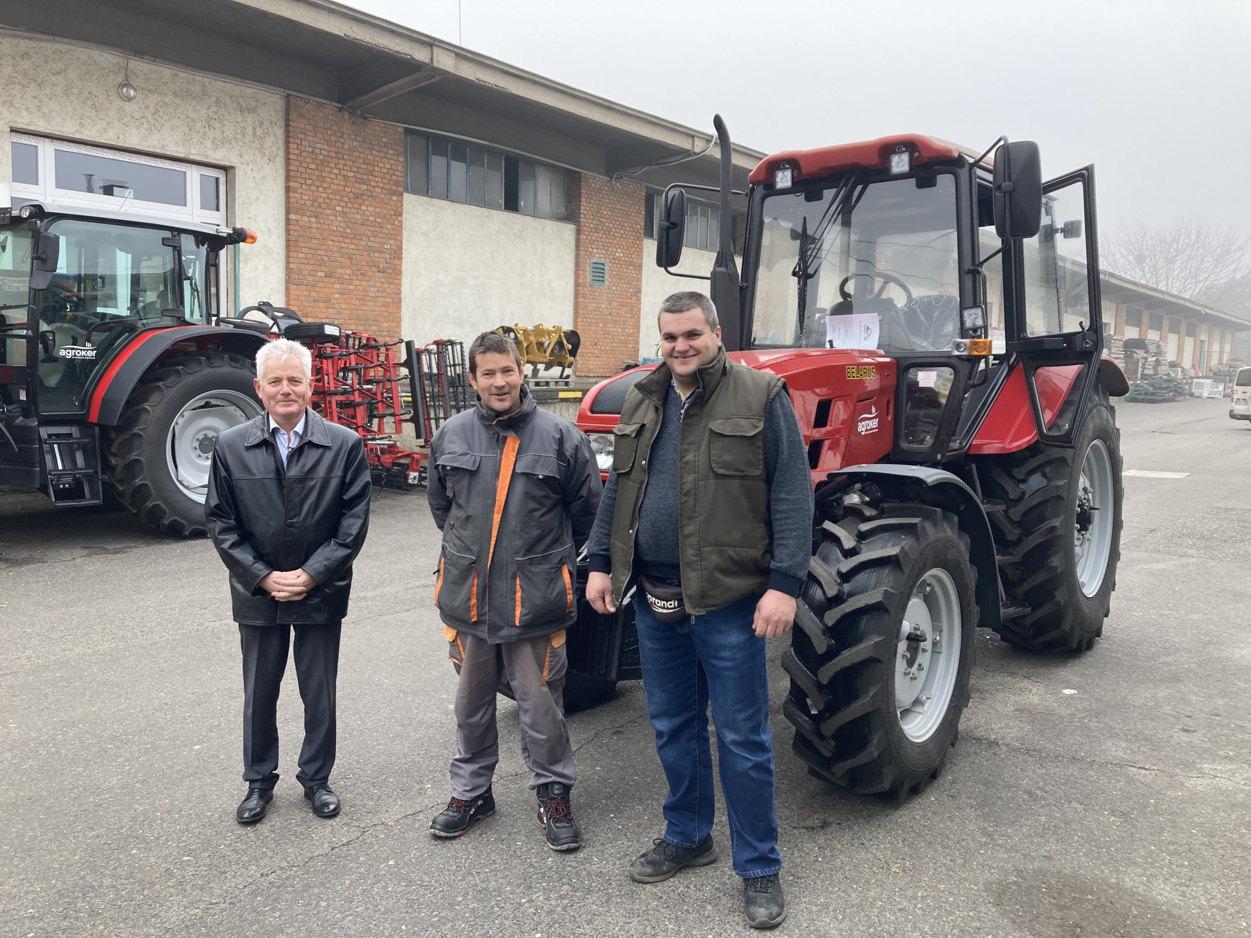 Három férfi állt egy piros traktor előtt, egy ipari épület mellett. A bal oldali férfi hivatalos öltözetben fekete kabátban, a középső munkáskabátban és nadrágban, a jobb oldali pedig mellényben és farmerben. Egy másik traktor látható a háttérben.