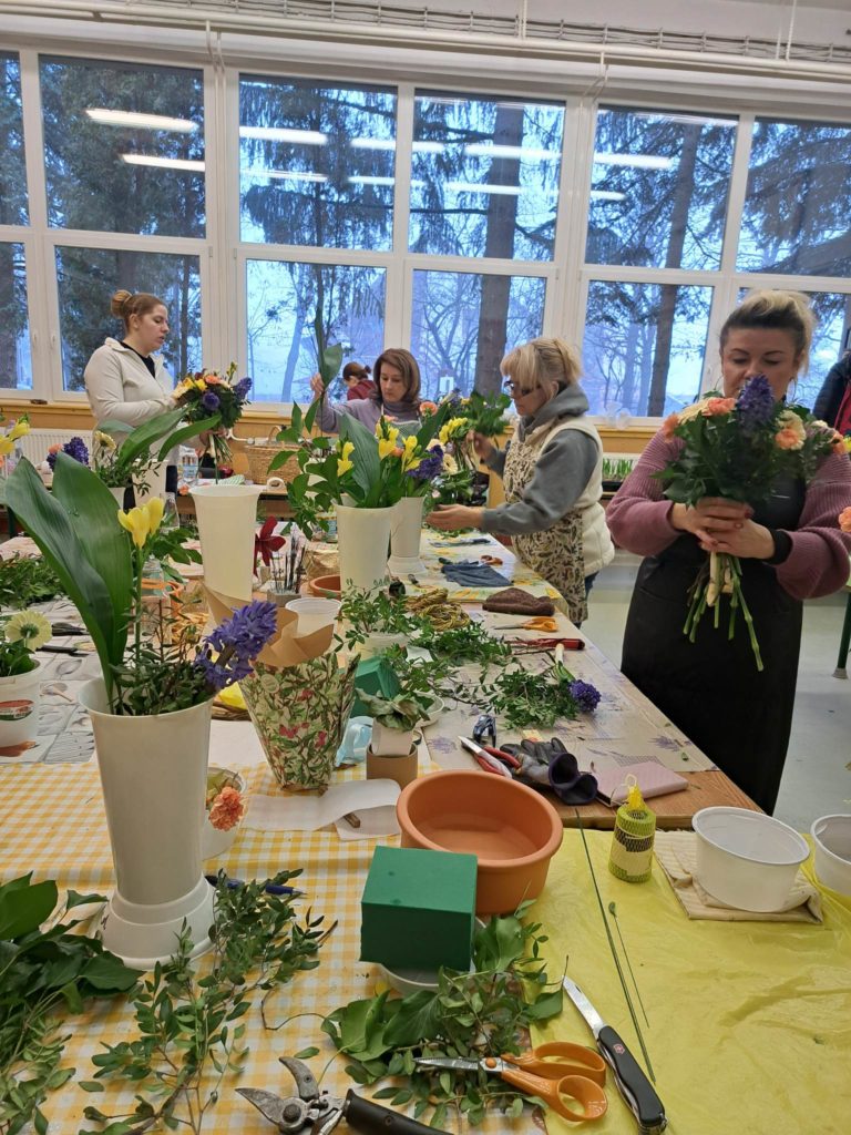 Emberek csoportja, akik színes virágokat rendeznek egy műhelyben. Az asztalokat különféle virágok, levelek és vázák borítják. A háttérben lévő nagy ablakok szabadtéri jelenetet mutatnak be fákkal és ködös légkörrel. A résztvevők koncentráltak és elkötelezettek kreatív feladataikban.