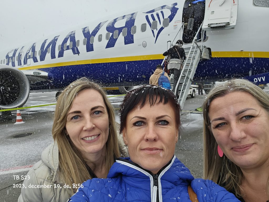 Három nő szelfit készít a Ryanair repülőgépe előtt egy havas napon, és részben látható a gép neve. Mindannyian meleg ruhát viselnek, és más utasok is láthatók, amint a mögöttük lévő lépcsőn felszállnak a gépre. A dátum „2023, december 10” látható.