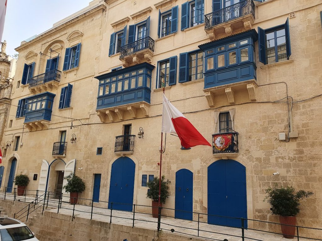 Bézs színű kőépület kék ajtókkal és redőnnyel, valamint több zárt kék erkéllyel. Az épület elejére piros-fehér zászlót szereltek, az ajtókat pedig két cserepes növény szegélyezi. Az épület díszes erkélyekkel és emblémával rendelkezik a zászló mellett.