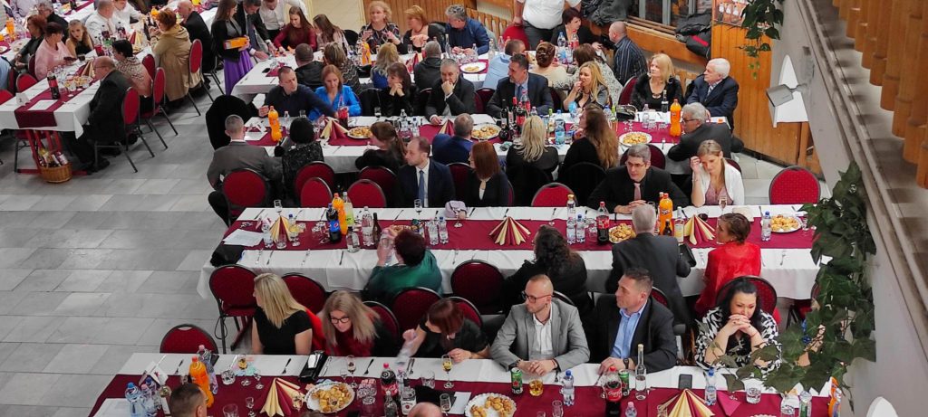 Nagy beltéri étkezési rendezvény több hosszú asztallal, melyeket vörös és fehér terítők díszítenek. A hivatalos ruhába öltözött résztvevők ülnek, beszélgetnek és élvezik az étkezést. Az asztalokon italos üvegek láthatók, beleértve a szódát és a vizet.