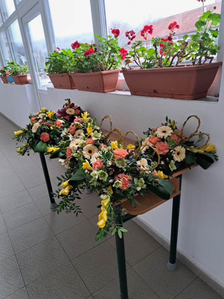 Az ablakpárkányra terített asztalon színes virágok és zöldek keverékével díszített kosaras virágdíszek láthatók. Az ablakpárkányon cserepes muskátli növények láthatók élénk piros és rózsaszín virágokkal egy jól megvilágított szobában.