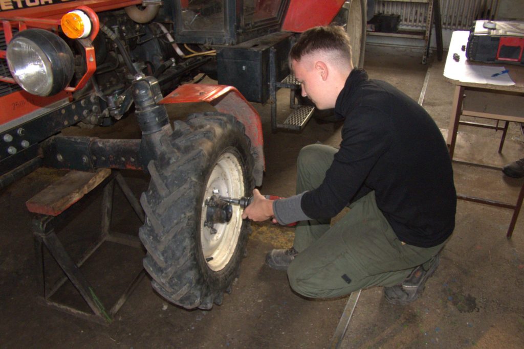 Egy fekete kapucnis pulcsit és zöld nadrágot viselő személy elektromos szerszámmal dolgozik egy piros traktor első kerekén egy műhelyben. A traktort faállványra emelik, a környéken különféle szerszámok, felszerelések vannak szétszórva.
