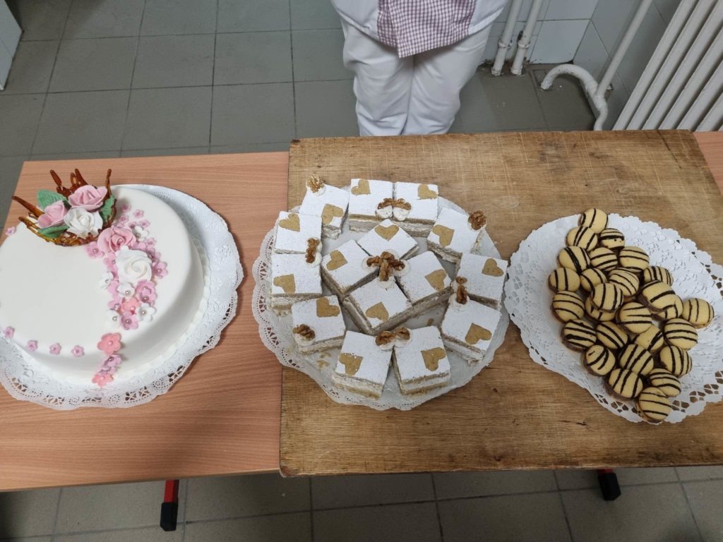 Egy asztal három különböző desszerttel: egy kerek fehér torta, rózsaszín virágokkal és levelekkel díszítve egy szalvétán, egy téglalap alakú, négyzet alakú tortatálca fehér cukormázzal és arany díszítéssel, valamint egy tányér kerek, csíkos sütivel. Egy személy áll a háttérben.