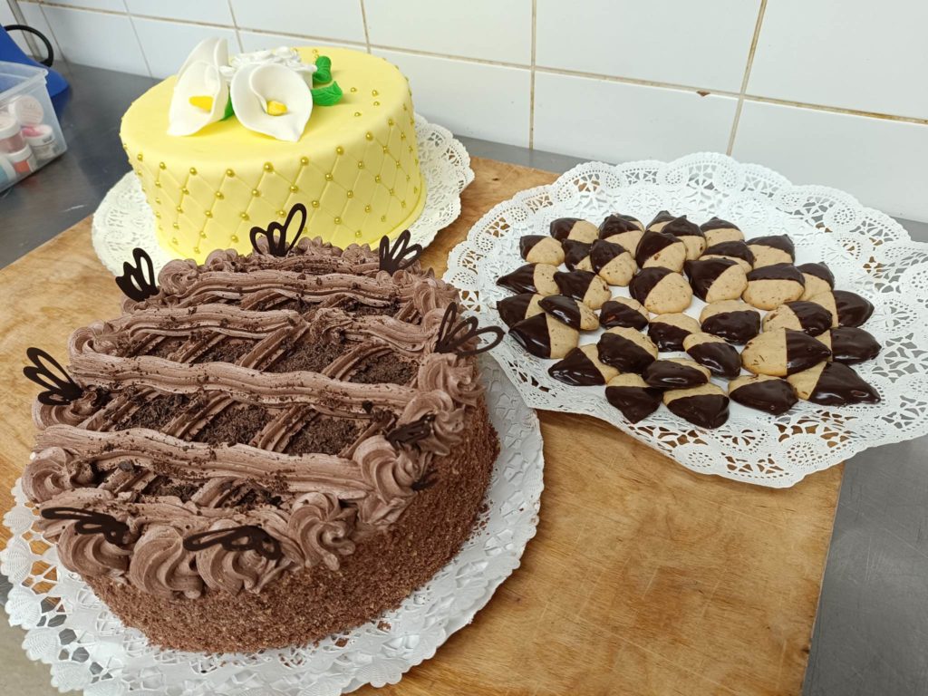 Két sütemény és egy tálca süti látható egy fa felületen. Az első torta sárga cukormázas fehér virágdíszekkel. A második torta csokoládé csokoládé pillangós díszítéssel. A sütiket részben csokoládéba mártjuk, dekoratívan elrendezzük.
