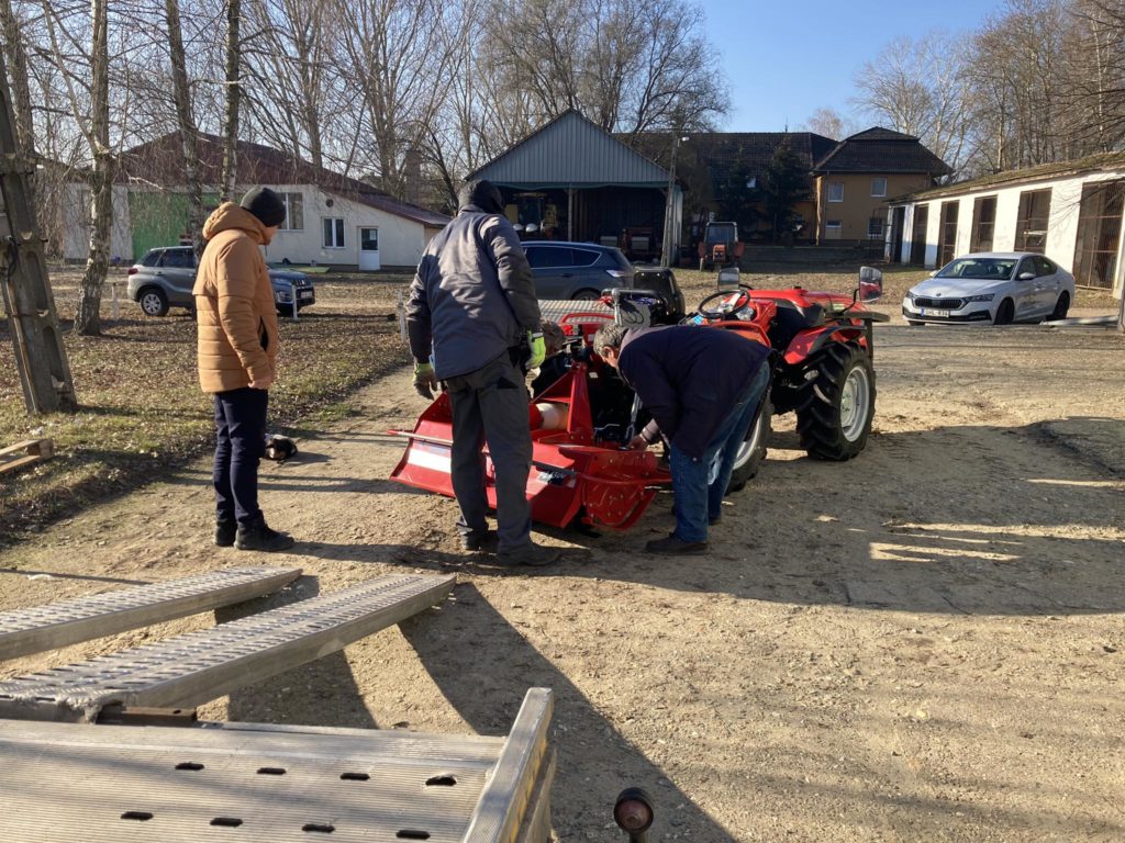 Három téli ruhába öltözött ember dolgozik egy piros mezőgazdasági gépen a szabadban egy napsütéses napon. Egy piros traktor van a géphez kapcsolva. A háttérben autók és épületek láthatók, csupasz fák pedig hideg évszakot jeleznek.