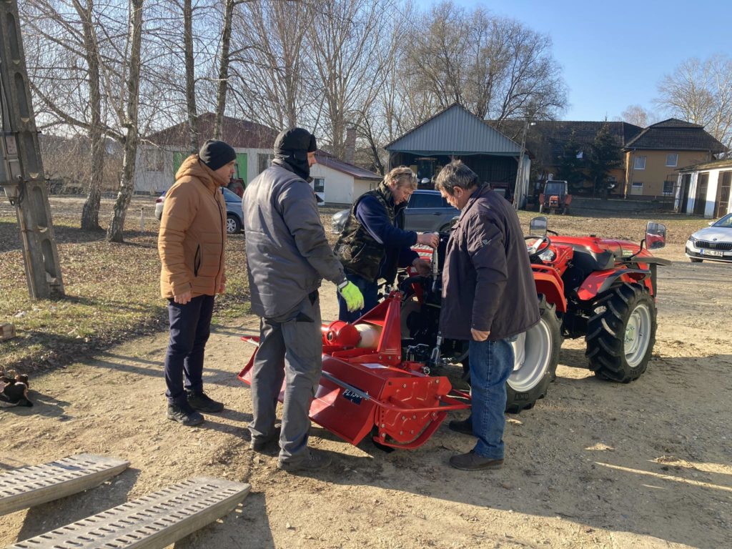 Négy személy áll körül, és egy piros traktorra erősített mezőgazdasági gépet vizsgálnak. Egy napsütéses napon a szabadban vannak, a háttérben épületek, fák és járművek láthatók. Úgy tűnik, hogy a csoport vitában vagy ellenőrzésben vesz részt.