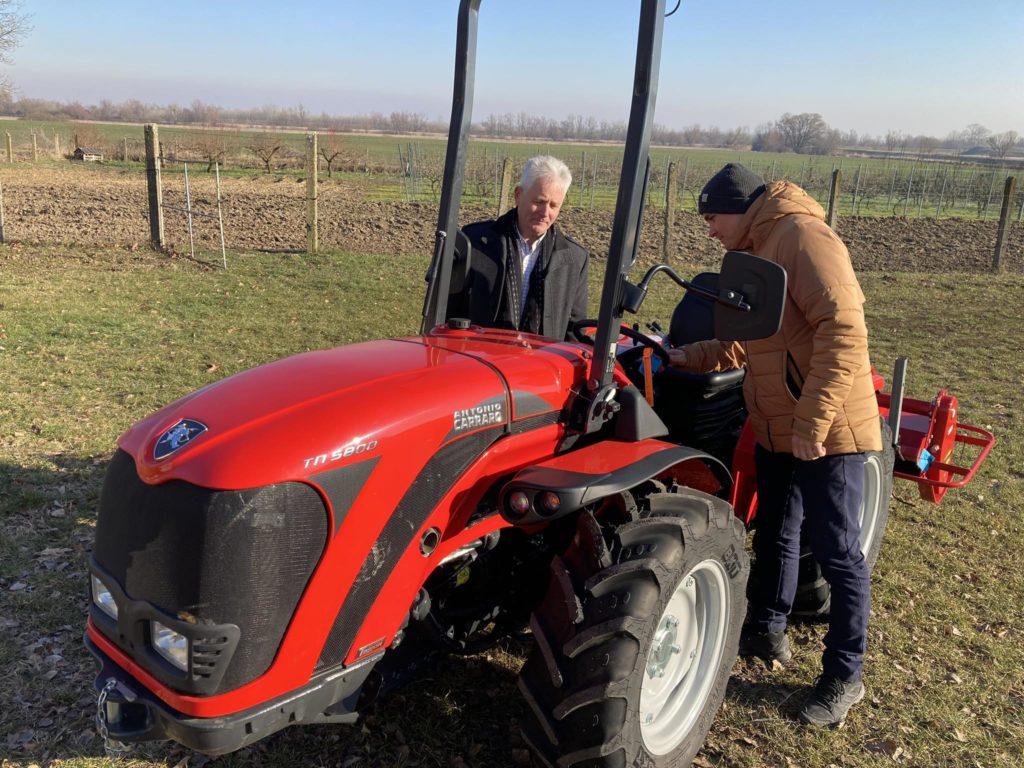 Két férfi áll egy piros traktor mellett egy vidéki mezőn. Az egyik férfi bézs kabátban és sapkában aktívan bánik a traktor kezelőszerveivel, míg a másik öltönyös érdeklődve figyel. A háttérben tiszta égbolt és bekerített termőföld látható.