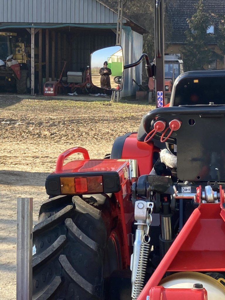 Közeli kép egy piros traktor hátuljáról egy farmon. A traktor egy koszos felületen parkol egy raktárépület közelében. A traktoron lévő oldalsó tükör egy álló, telefonnal fényképező embert tükröz. Különféle mezőgazdasági berendezések láthatók a háttérben.