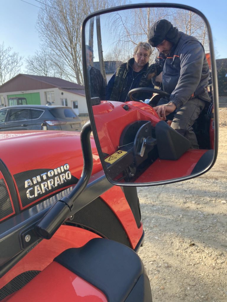 Két ember dolgozik egy piros Antonio Carraro traktoron. Az egyik sapkát és téli ruhát viselő személy állít valamit a traktor kezelőszervei közelében, míg a másik megfigyel. A jelenet egy oldaltükörben tükröződik, a háttérben házak és autók.