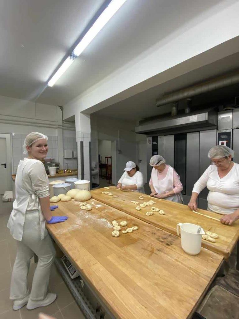 Egy négyfős, fehér egyenruhát és hajhálót viselő nőből álló csoport dolgozik egy pékségben. Egy nő a bal oldalon mosolyog a kamerába, miközben egy tésztás asztal mellett áll. A másik három nő tésztát formáz a nagy faasztalnál. Ipari sütők állnak a háttérben.