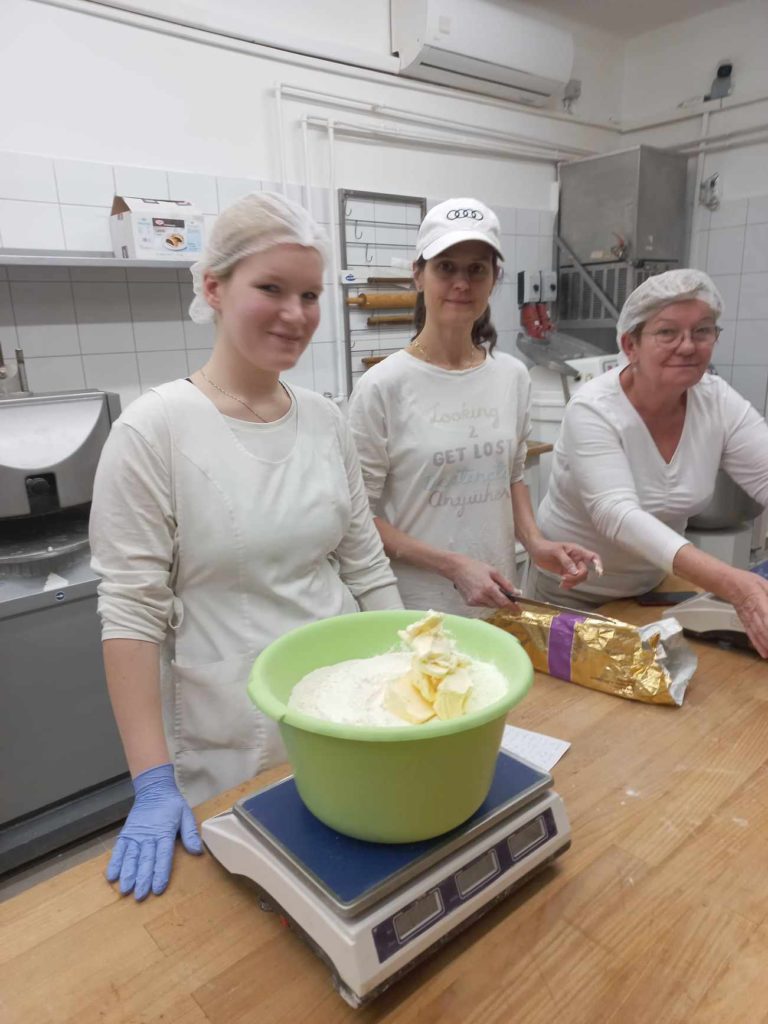 Három nő dolgozik egy konyhában. A bal oldali nő hajhálót, kesztyűt és fehér inget visel. Középen egy fehér sapkás és inges nő áll a mérlegen egy nagy tál liszttel és vajjal. A jobb oldali nő vajat vág.
