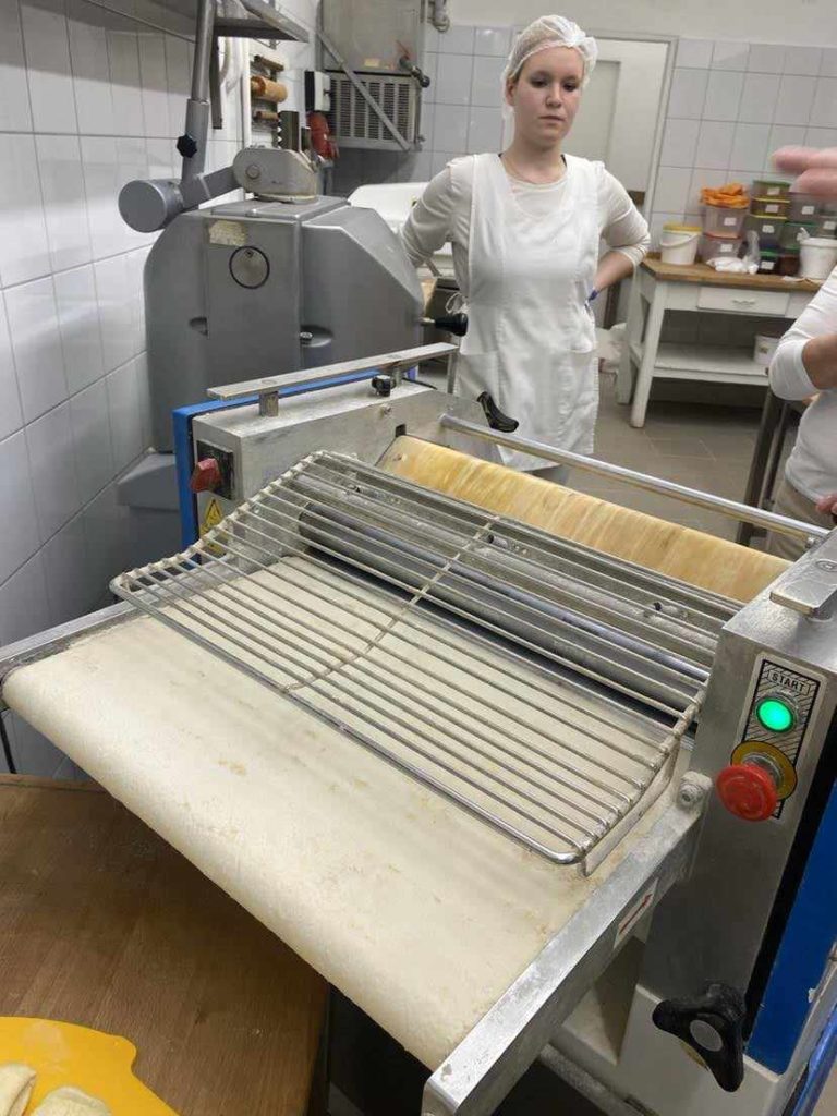 Egy fehér egyenruhás, hajhálós nő áll egy tésztagép mellett a konyhában. A gép használatban van, vékony lapokra sodorja a tésztát. A háttérben a konyhai kellékekkel és hozzávalókkal ellátott polcok láthatók.
