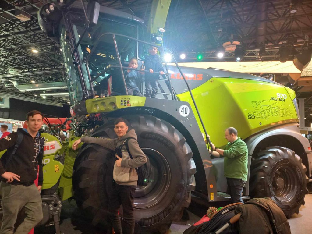 Egy csoport ember egy nagy zöld mezőgazdasági gép körül gyűlik össze, „CLAAS” márkajelzéssel. Úgy tűnik, hogy a jelenet bent van, valószínűleg egy rendezvényen vagy kiállításon. Két személy ül a gép kabinjában, míg mások mellette állnak, mosolyogva a kamerába.