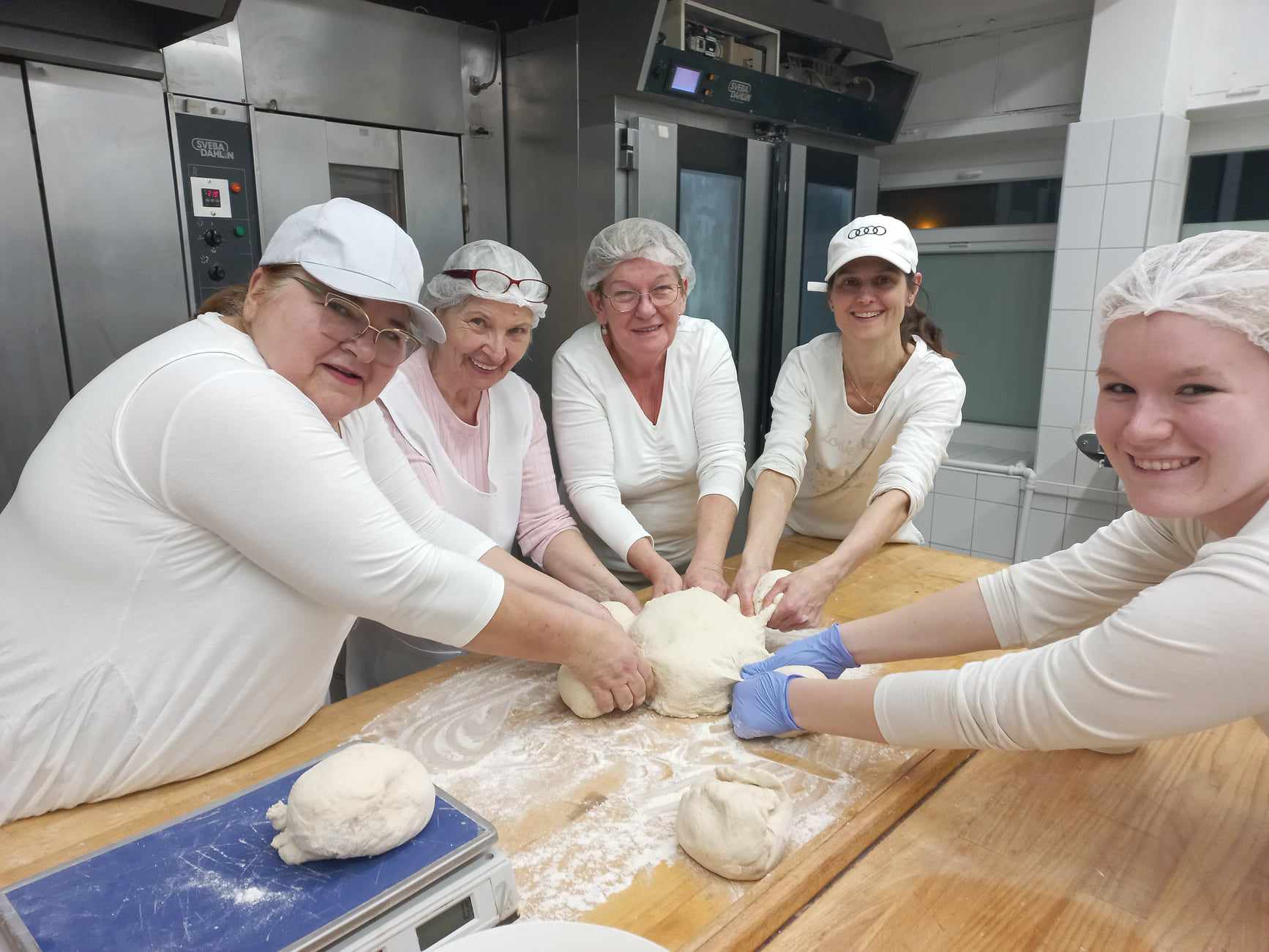 Egy ötfős nőből álló csoport, akik mindegyike fehér inget és hajtakarót visel, mosolyogva dolgoznak együtt, hogy tésztát gyúrjanak egy nagy faasztalon egy pékség konyhájában. A konyha tele van sütőberendezéssel és sütővel a háttérben.