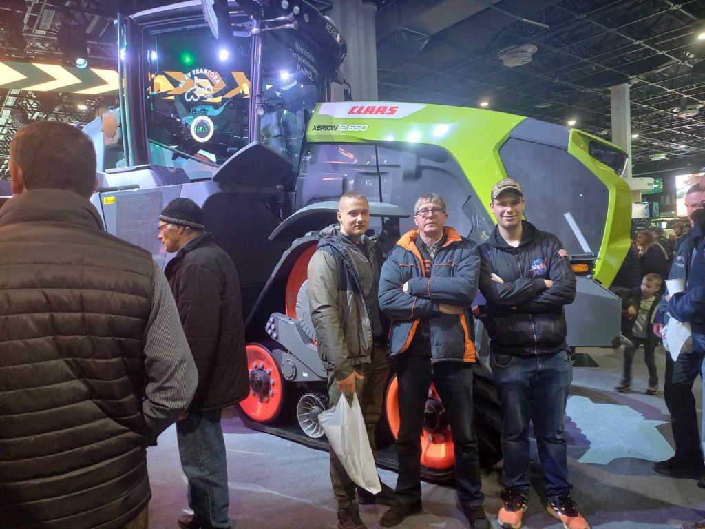 Három férfi pózol egy nagy zöld-fekete Claas Xerion 4500/5000 traktor előtt egy beltéri rendezvényen. A háttérben mások is láthatók, akik a traktort és a kiállítást nézik.