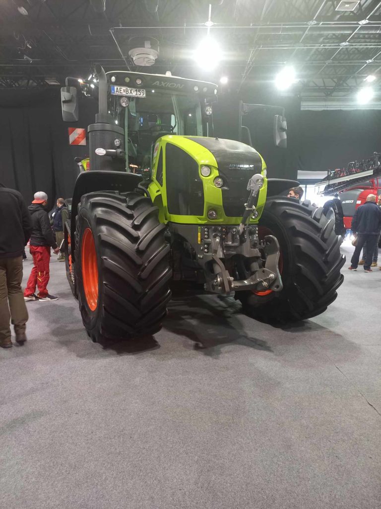 Egy nagy zöld-fekete traktor masszív gumiabroncsokkal van kiállítva egy kiállításon vagy kongresszuson. A háttérben többen figyelik a traktort és más gépeket. A teret erős mennyezeti lámpák világítják meg.