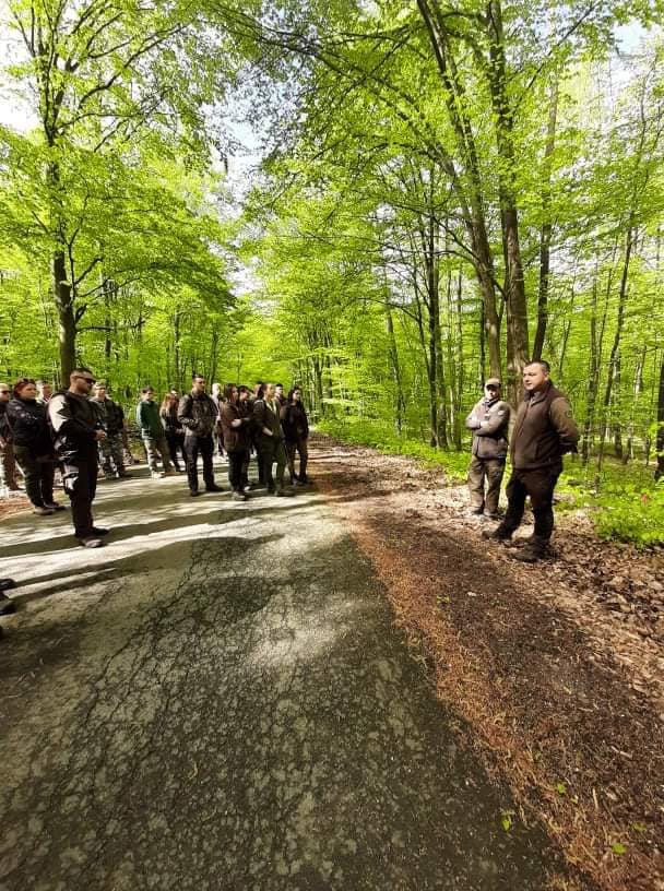 Egy csoport ember egy ösvényen áll egy buja, zöld erdőben. Vannak, akik figyelmesen hallgatnak egy jobboldali férfit, aki úgy tűnik, beszél vagy utasításokat ad. A napfény átszűrődik a fákon, és meleg ragyogással világítja meg a jelenetet.