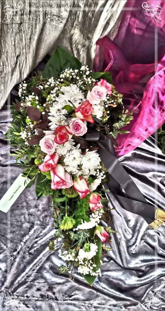 Virágkompozíció rózsaszín és fehér rózsa, fehér krizantém, baba lehelet és zöld lomb keverékével. A csokor fekete szalaggal van körbekötve, és szürke textúrájú szöveten nyugszik, háttérben bíbor szövettel.