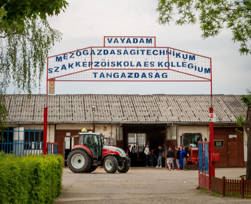 Egy traktor parkol egy mezőgazdasági technikum épülete előtt, íves táblával a feje fölött. A táblán ez áll: "Vay Ádám Mezőgazdasági Technikum Szakképző Iskola és Kollégium Tangazdaság." Több ember gyűlt össze az épület bejáratánál.