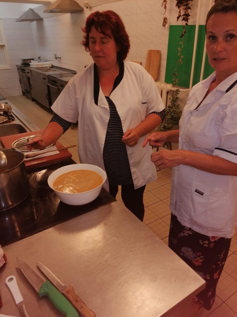 Két fehér konyhai egyenruhát viselő nő egy profi konyhában tartózkodik. Egy nő habverőt tart egy sárga keverékkel töltött keverőtál fölött. A másik nő a tálra mutat. A háttérben különféle edények és konyhai felszerelések láthatók.