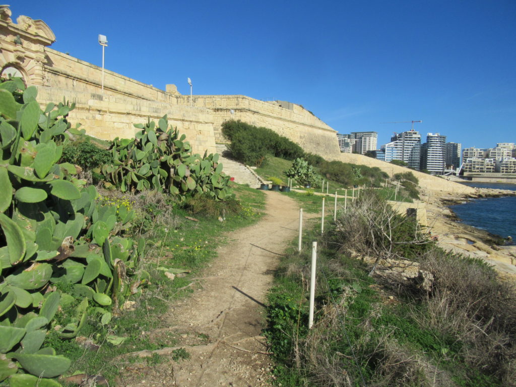 A part mentén földút vezet, balról kaktuszok és zöld cserjék szegélyezik, balról szintén kőerődfal. Modern épületek láthatók a távolban a jobb oldalon, a kék tenger mellett, tiszta égbolt alatt.
