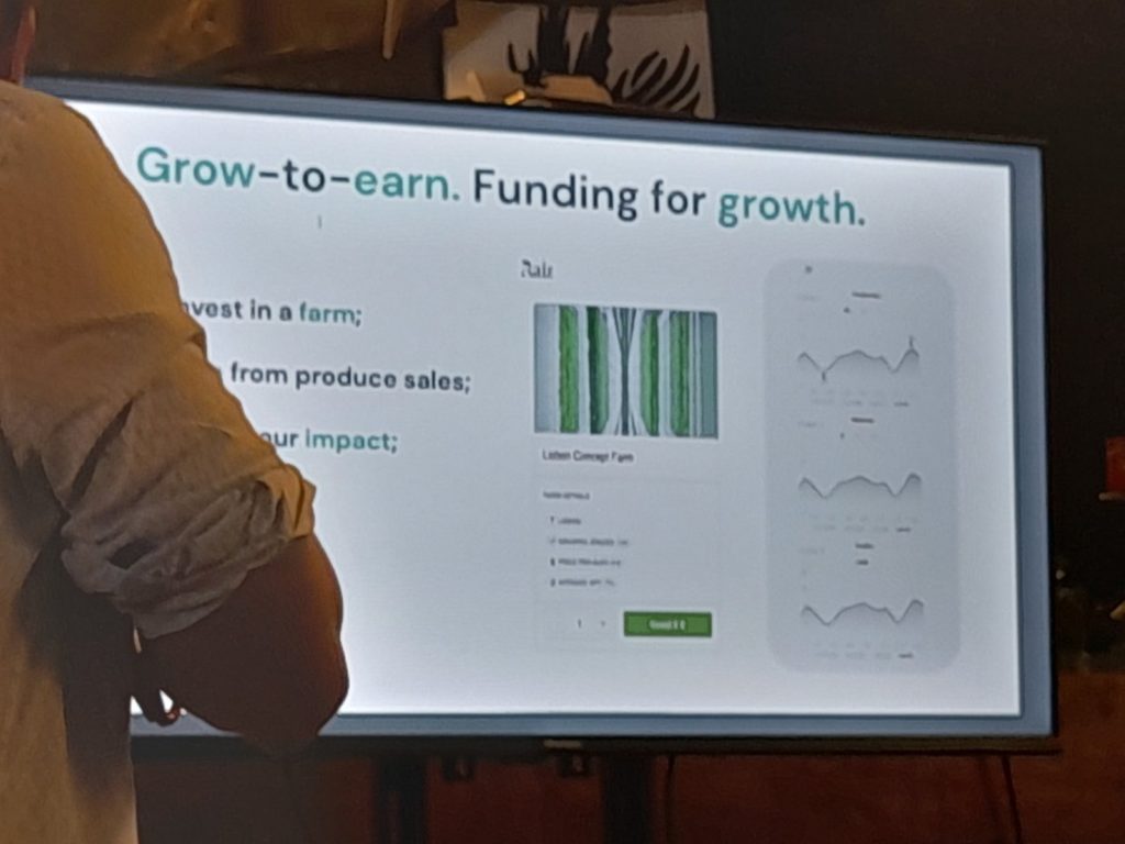 Egy személy egy prezentációs diát megjelenítő nagy képernyő előtt áll. A dia a "növekedés a keresethez" és a növekedés finanszírozásával foglalkozik, bemutatja a gazdaságokba történő befektetéseket, a termékértékesítésből származó bevételeket és a hatás nyomon követését. A telefon képernyője is látható a kapcsolódó adatokkal.