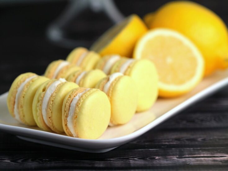 Egy téglalap alakú fehér tányéron citromízű macaronok sorakoznak el. A macaronok világossárgák, krémes töltelékkel. A háttérben egész és félbevágott citromok láthatók, amelyek friss citrusos hatást adnak a jelenethez.