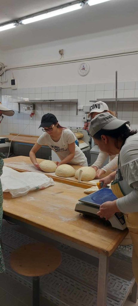 Három személy egy profi konyhában kenyértésztát formáz egy fapulton. Az előtérben egy ember kötényt visel, és mérlegen méri a tésztát. A szoba falai fehér csempézettek, háttérben konyhai felszerelések.