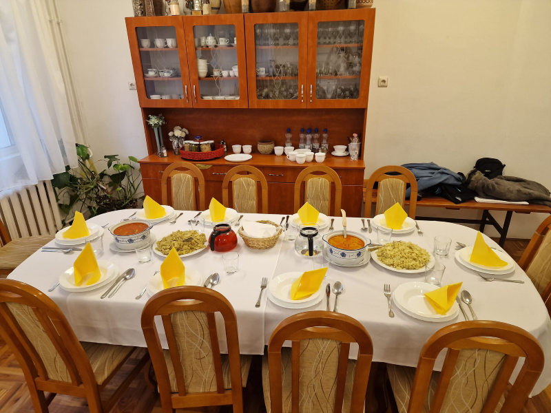 Nyolc személyes étkezőasztal fehér ruhával, sárga szalvétákkal, tányérokkal és evőeszközökkel. Az asztalon több tál étel, egy piros tégely és egy szőtt kenyérkosár található. Az asztal mögött egy fából készült porcelán szekrényben edények és üvegáru látható. Növények díszítik a sarkot.