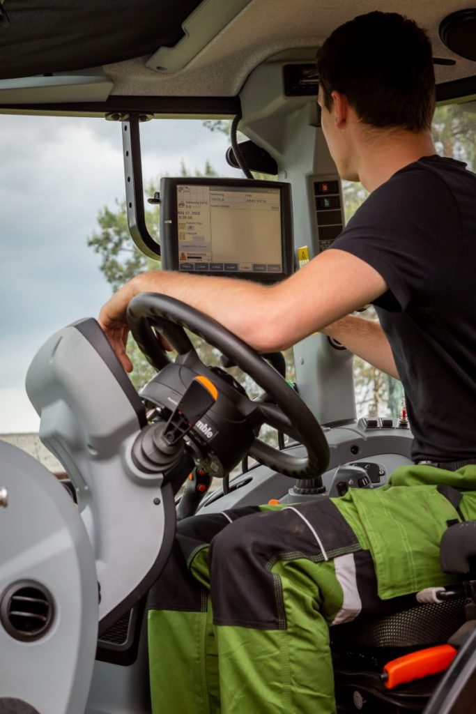 Egy zöldnadrágos és fekete inges személy traktort vagy nehézgépet kezel, a fülkében lévő érintőképernyős monitorra összpontosítva. Az egyén beállítja a kezelőszerveket, és az ablakon keresztül látszanak a fák.