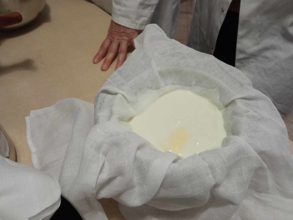 Egy fehér köpenyes ember tejet aludt egy tálban egy fehér sajtkendővel. A sajtkendőt a tálra terítik, és úgy tűnik, hogy a tej besűrűsödik, és túróvá válik, valószínűleg a sajtkészítési folyamat részeként. Az egyik kéz a pulton nyugszik.