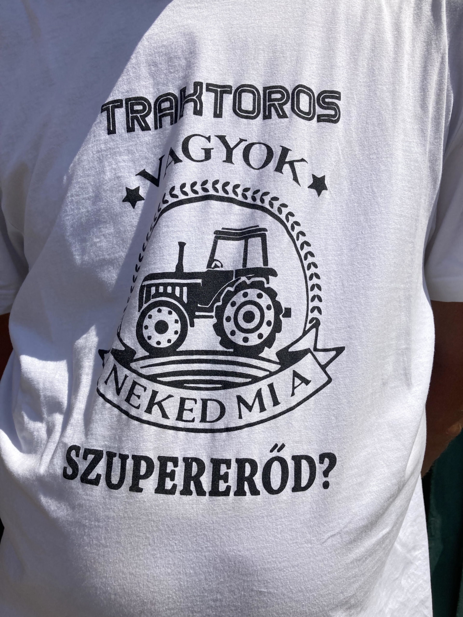 Egy személy fehér pólót visel, magyar nyelvű grafikai tervezéssel és szöveggel. A kivitelben egy traktor képe látható. A szöveg így szól: "TRAKTOROS VAGYOK NEKED MIA SZUPERERŐD?" ami azt jelenti: "Traktoros vagyok, mi a szuperképességed?" angolul.