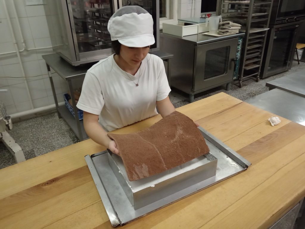 Egy fehér inget és sapkát viselő személyt látnak egy pékségben, aki egy nagy csokitortát helyez egy fehér krémmel töltött téglalap alakú forma fölé. A háttérben több konyhai berendezés látható.