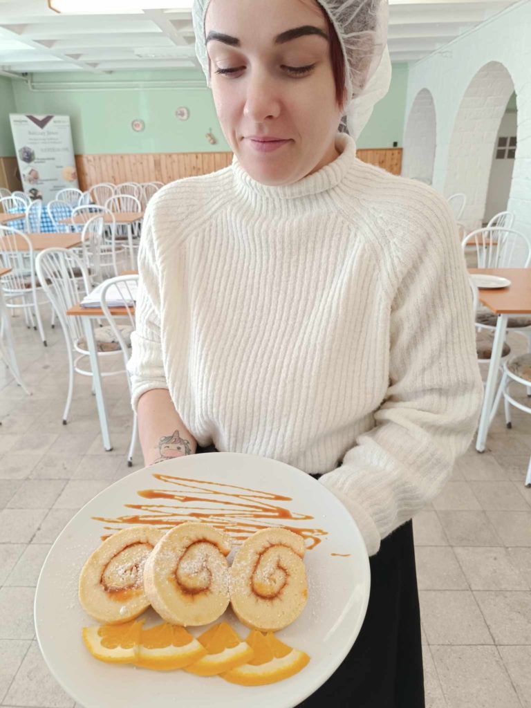 Egy fehér pulóvert és hajhálót viselő nő egy fehér tányért tart kezében, amelyen három, porcukorral meghintett, karamellszósszal meglocsolt és narancsszeletekkel díszített péksütemény található. Egy fénnyel teli étkezőben áll, fehér székekkel és asztalokkal a háttérben.