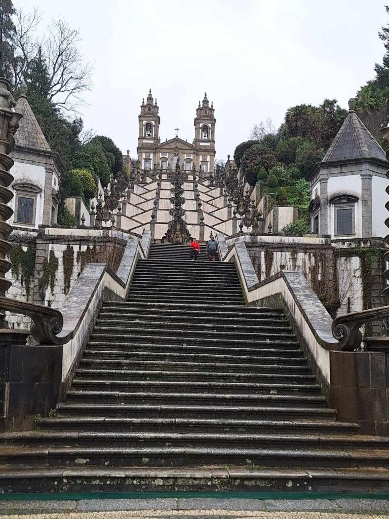 Szimmetrikus kőlépcsőkkel és dekoratív korlátokkal rendelkező, hatalmas lépcső vezet fel egy barokk stílusú, kéttornyú templomhoz, amelyet buja növényzet vesz körül. Néhány ember látható a lépcsőkön, és ez egyfajta léptéket ad az impozáns építészethez.