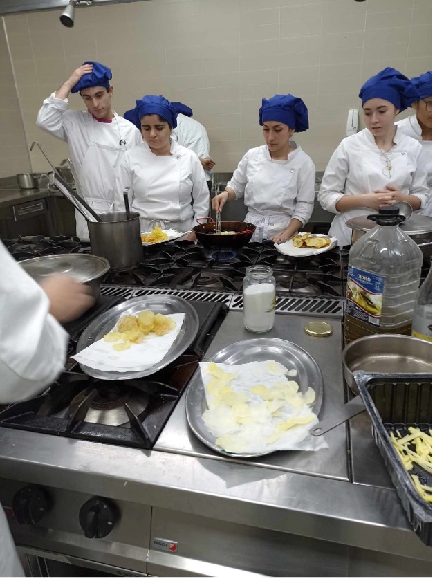 Fehér egyenruhát és kék sapkát viselő diákok főznek egy profi konyhában. Az egyik diák ételt kever egy serpenyőben, míg mások sültnek tűnő ételeket készítenek a tányérokon. A főzőedényeket és a hozzávalókat szétszórják a főzőlapokon.