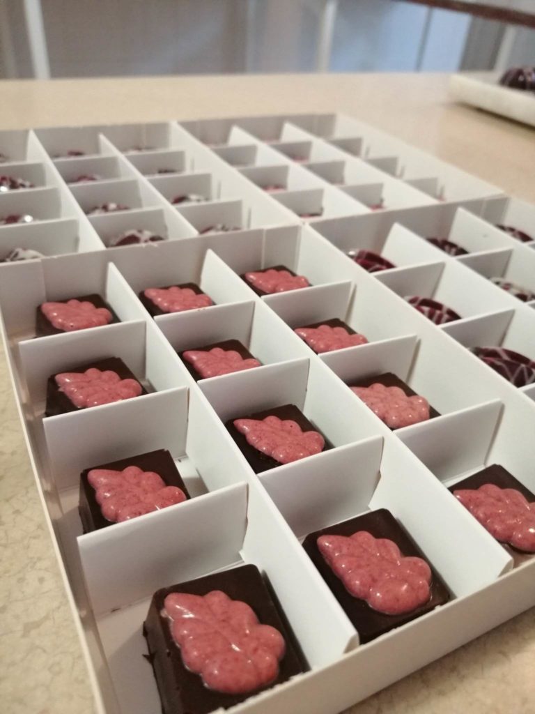 Közeli kép egy fehér dobozról, amelyben különböző négyzet alakú csokoládék vannak rózsaszín, virág alakú feltétekkel. A csokoládék szépen elrendezve az egyes rekeszekben. A háttér enyhén elmosódott, a csokoládékra irányítva a figyelmet.