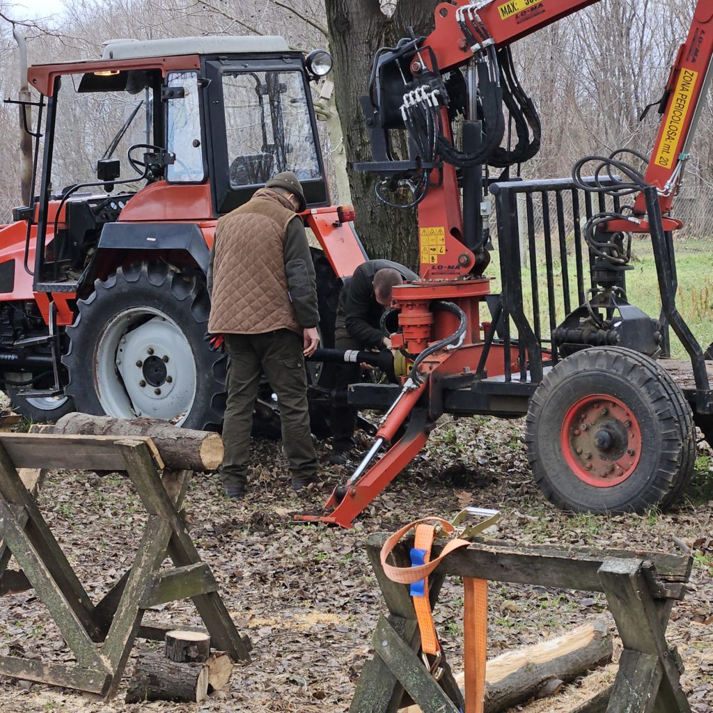 Két ember – egyikük barna, másikuk fekete kabátban – egy traktorra erősített nagy piros rönkhasító gépet üzemeltet egy erdős területen. A rönkök egy fém állványon vannak a közelben. A terület sáros, lehullott levelekkel.