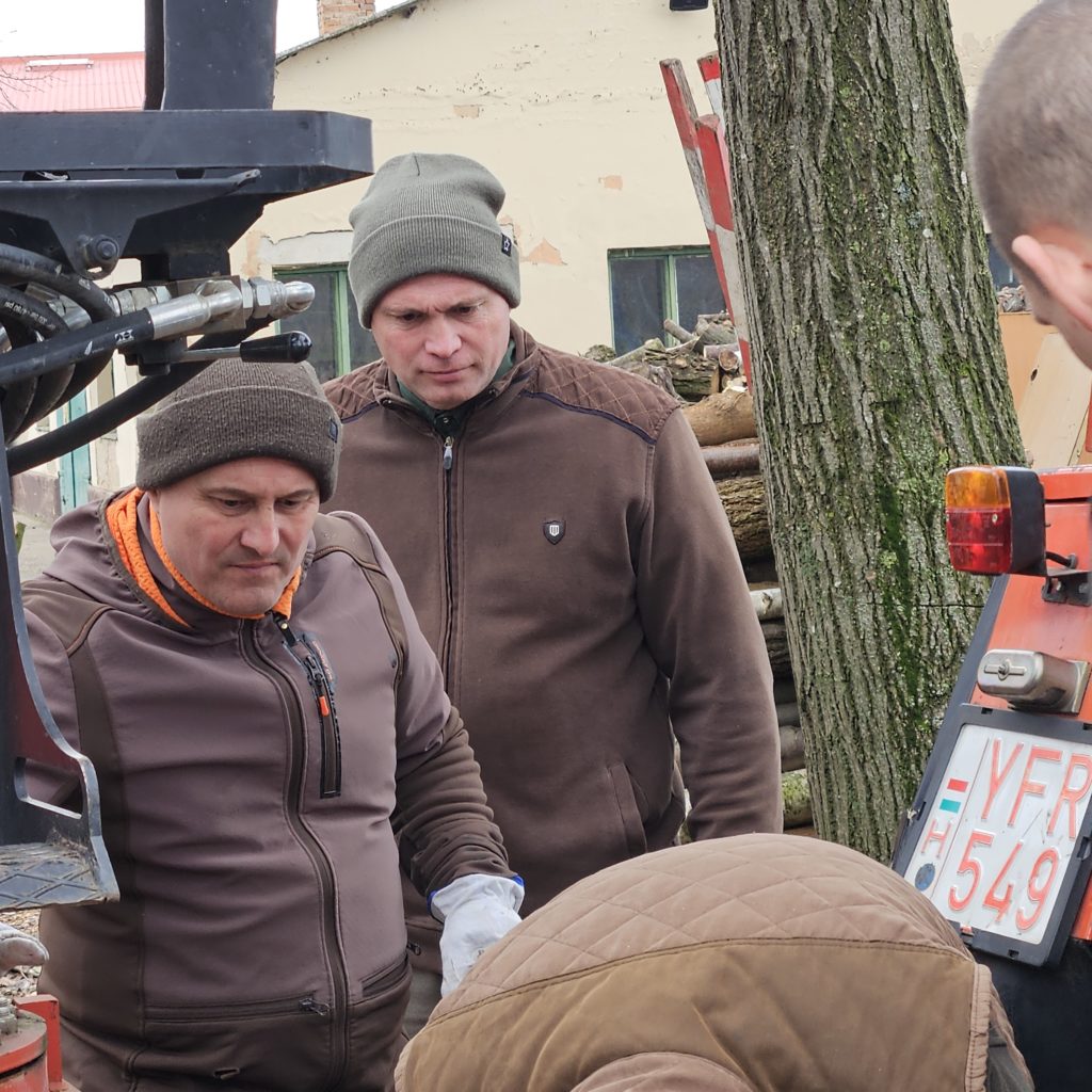 Két barna kabátba és kabátba öltözött férfi áll a szabadban egy traktor mellett, és gépeket üzemeltet. Az egyik férfi figyelmesen nézi a berendezést, míg a másik kissé hátrébb áll. A háttérben egy fa és egy ipari épület látható.