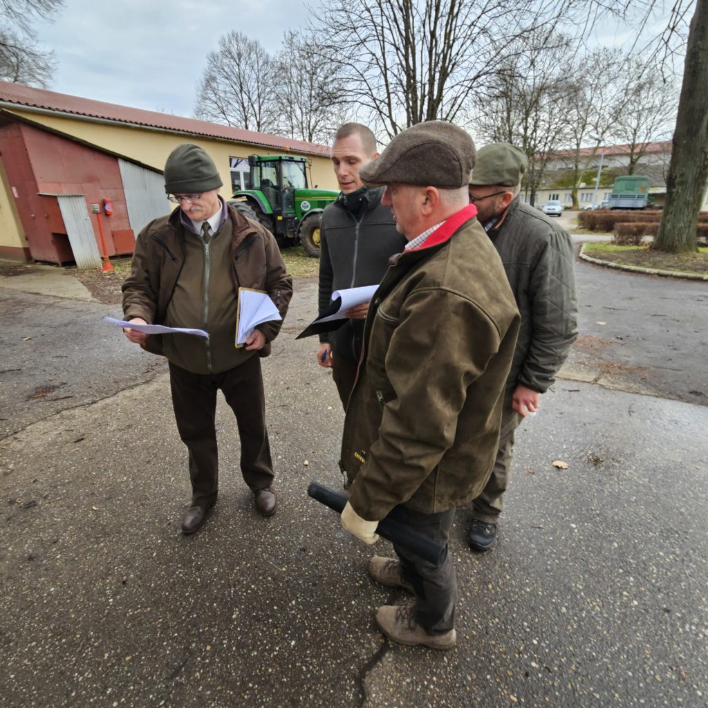 Négy kültéri ruhás ember áll egy gazdasági épület közelében, és papírokat nézeget. Egy személy a dokumentumokra mutat, míg mások nézik. Egy zöld traktor látható a háttérben, és úgy tűnik, hogy a jelenet egy vidéki területen játszódik.