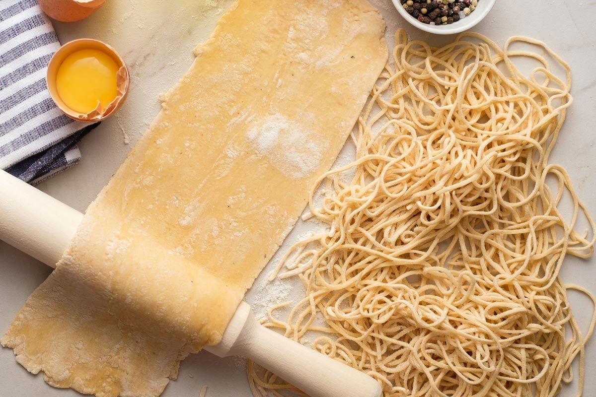 Kinyújtott tészta tésztalap márvány felületen sodrófa mellett, feltört tojáshéj, tojássárgája, csíkos kendő és egy tál bors. A kép jobb oldalán egy halom frissen vágott spagetti látható.