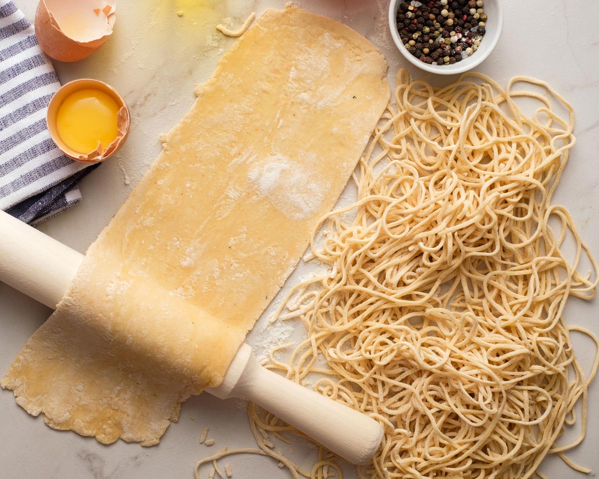 Kinyújtott tészta tésztalap márvány felületen sodrófa mellett, feltört tojáshéj, tojássárgája, csíkos kendő és egy tál bors. A kép jobb oldalán egy halom frissen vágott spagetti látható.