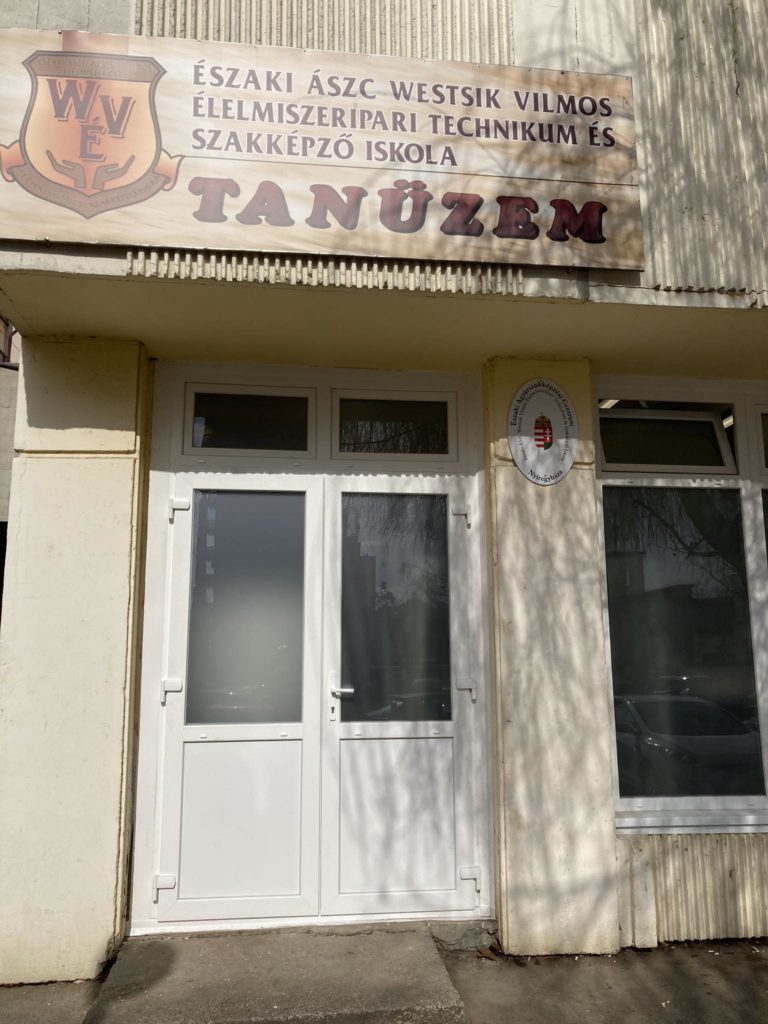 Épülethomlokzat magyar felirattal, amelyen az "Északi ÁSZC Westik Vilmos Élelmiszeripari Technikum és Szakképző Iskola TANÜZEM" felirat olvasható. Mindkét oldalon két fehér ajtó és ablak található. Az épület világos, texturált külsővel rendelkezik.
