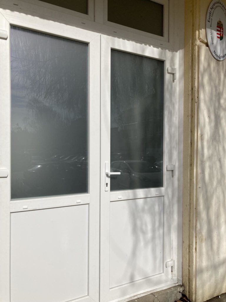 Egy épület külsején egy fehér dupla ajtó, mattüveg panelekkel, fehér keretben található. Az ajtótól jobbra egy kerek tábla, pajzsjelzéssel és körülötte szöveggel. Faágak árnyéka vetül az ajtóra.