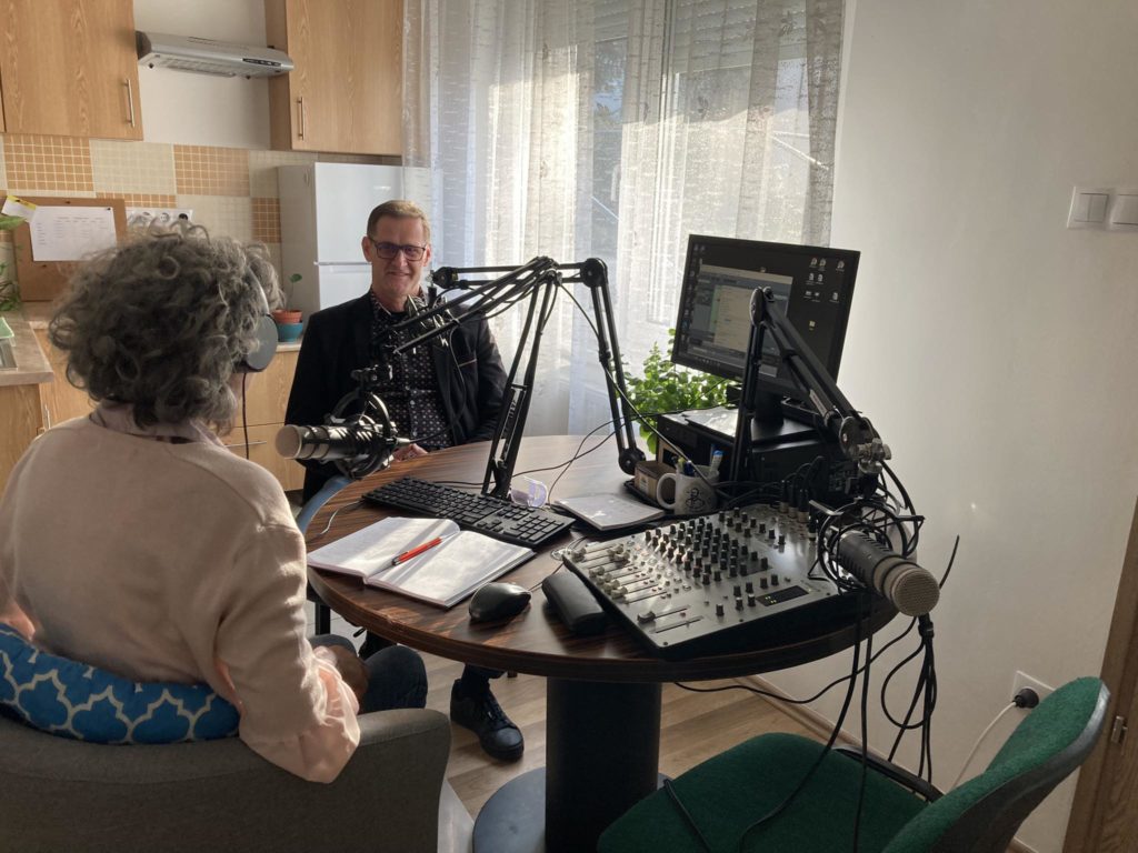 Két ember ül egymással szemben egy asztalnál egy otthoni stúdióban. Mindketten fejhallgatót viselnek, és mikrofonba beszélnek. Az asztalon számítógép, keverőtábla és dokumentumok találhatók. A háttérben lévő ablakon keresztül beszűrődik a napfény.