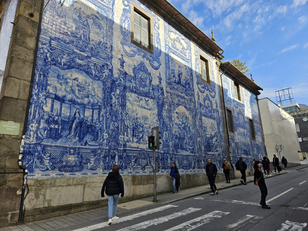 Egy nagy falfestmény részletgazdag kék-fehér csempével díszíti az épület oldalát. Emberek sétálnak a járdán és az utcákon a falfestmény előtt. Úgy tűnik, hogy a műalkotás történelmi vagy vallási jeleneteket ábrázol. A fenti égbolt többnyire derült.