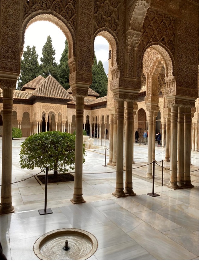 A képen a granadai Alhambrában található Oroszlánok udvara látható, bonyolultan faragott boltívekkel, márványoszlopokkal és egy központi szökőkúttal. Az udvart ápolt bokrok és fák szegélyezik, a környező hagyományos mór építészettel.